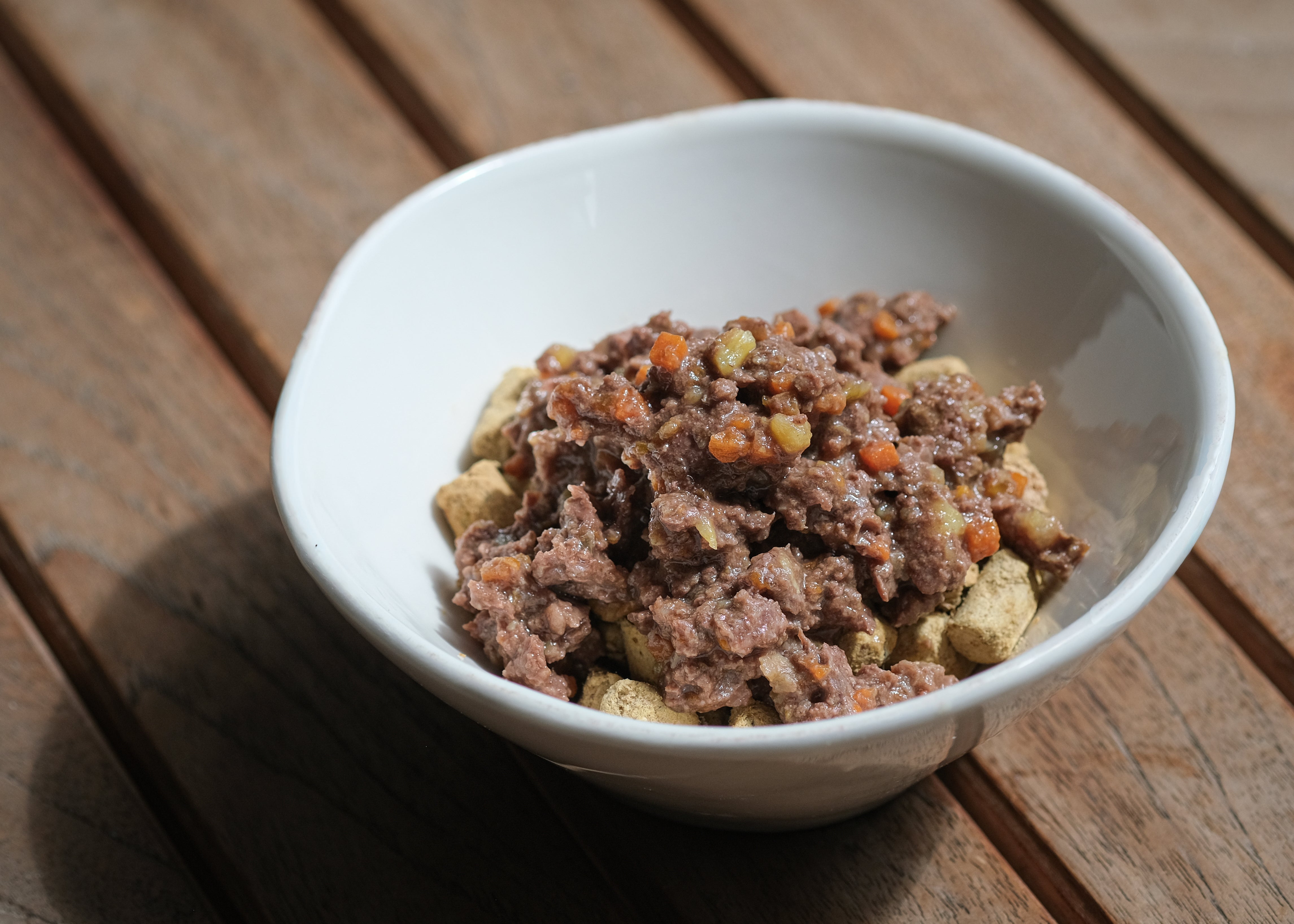 白い器に盛られたKaruizawa HarvestのVENISON & APPLEミールトッパー。茶色いドライフードの上に、にんじんなどの野菜を刻んで混ぜた赤みがかった鹿肉のトッピングがのっている。木製のテーブルの上に置かれている。